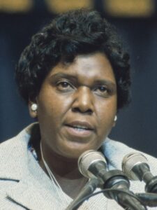 Barbara C Jordan wears white pearl earrings and a light colored blazer. She is speaking into two microphones