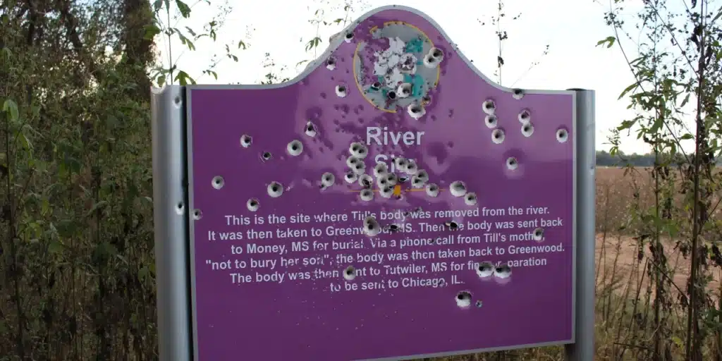 A photo of Emmett Till's Memorial marker by the river. It is riddled with bullet holes. 