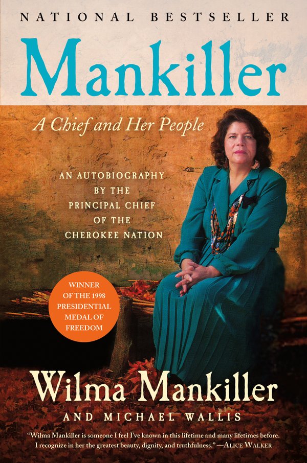 Wilma Mankiller is sitting on a bench, dressed all in a deep turquoise dress and blazer with a Native beadwork necklace.