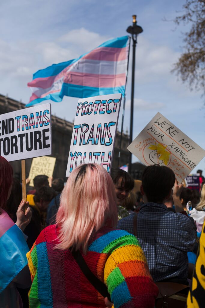 a group of people protest anti-trans legislation with signs that say "we're not your scapegoats", "protect trans youth" and "end trans torture"